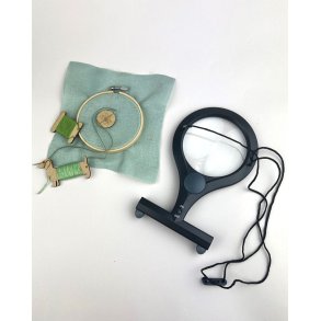 Svart handsfree f�rstoringsglas med LED-ljus och justerbar halsrem bredvid ett broderi.