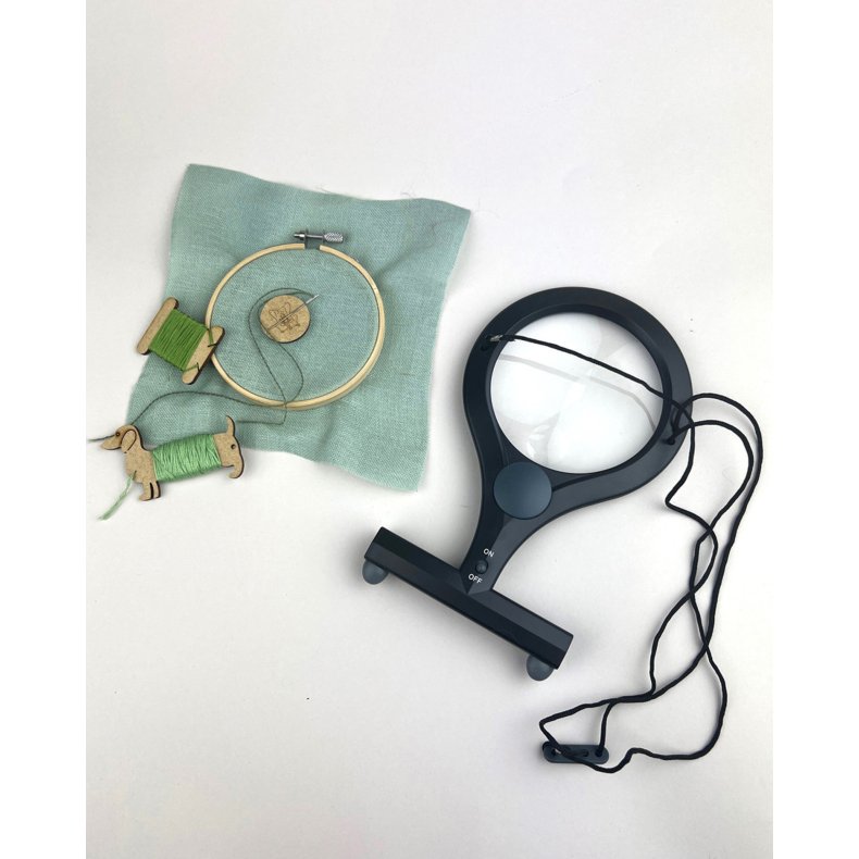 Svart handsfree f�rstoringsglas med LED-ljus och justerbar halsrem bredvid ett broderi.